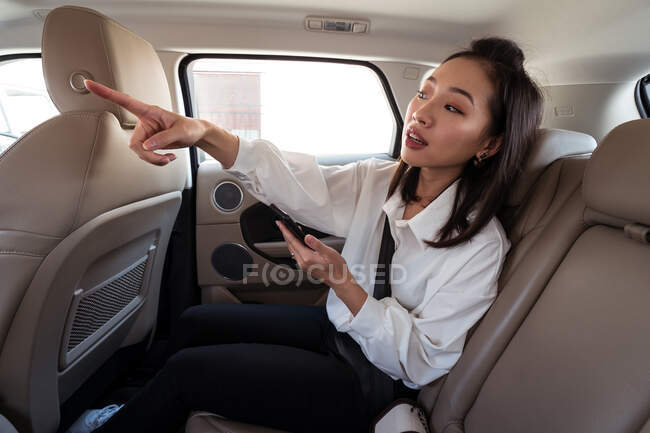 Vue latérale du passager ethnique féminin avec smartphone monté sur le siège arrière en taxi et montrant la direction du conducteur — Photo de stock