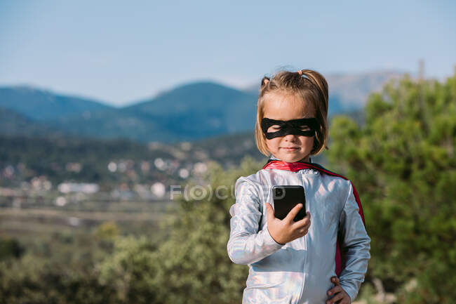 Self assured girl in eye mask superhero costume with cape browsing on cellphone — Stock Photo