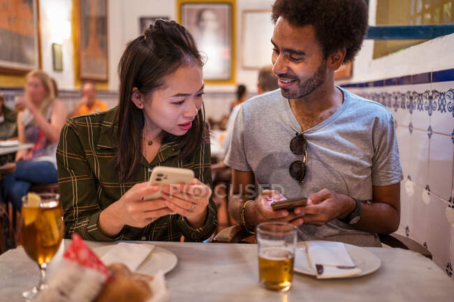 Щаслива молода багаторасова пара в повсякденному одязі, використовуючи смартфони, сидячи разом за столом з келихами пива в сучасному ресторані — стокове фото