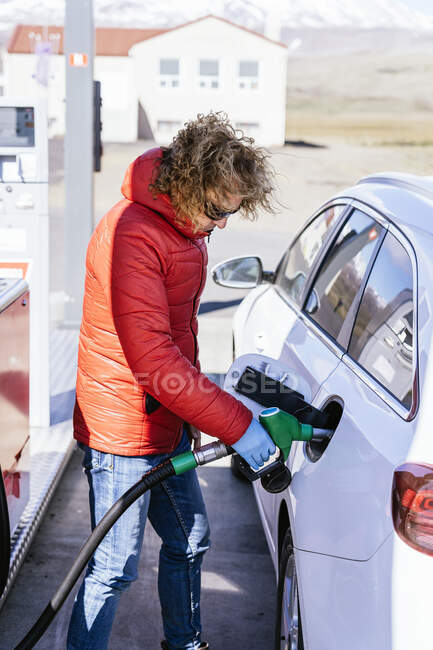 Visão lateral de um homem sério com cabelos encaracolados em roupas e luvas de látex em pé enquanto reabastece o carro no posto de gasolina em dia ensolarado — Fotografia de Stock