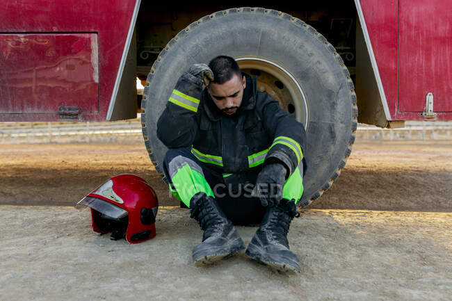 Estremamente maschio pompiere indossa uniforme seduto a terra vicino al motore del fuoco vicino al casco rosso mentre appoggia la testa a portata di mano — Foto stock