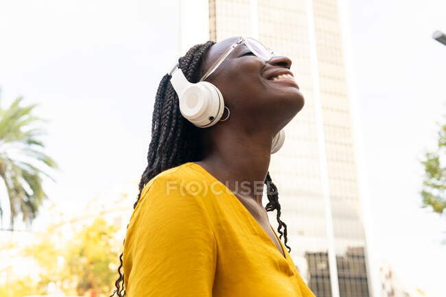 Vista lateral do meloman feminino afro-americano encantado ouvindo músicas em fones de ouvido enquanto estava na rua com edifício na cidade — Fotografia de Stock