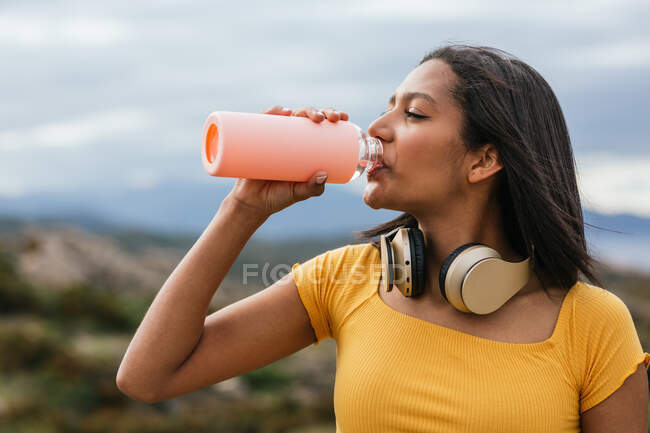 Mulher étnica sedenta com fones de ouvido sem fio no pescoço água potável de garrafa de plástico para refresco na natureza — Fotografia de Stock
