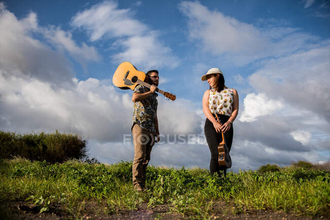 Baixo ângulo de músico homem alegre com guitarra acústica no ombro em pé na grama verde perto da fêmea com ukulele na natureza contra o céu azul no dia ensolarado — Fotografia de Stock
