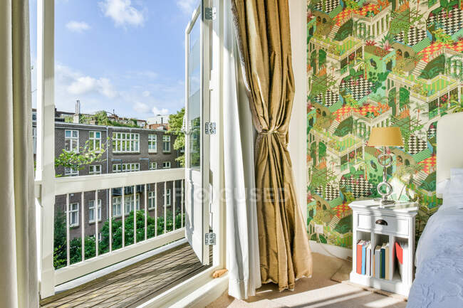 Открытые двери балкона с видом на город в просторной спальне с креативными обоями и белой мебелью в солнечный день — стоковое фото
