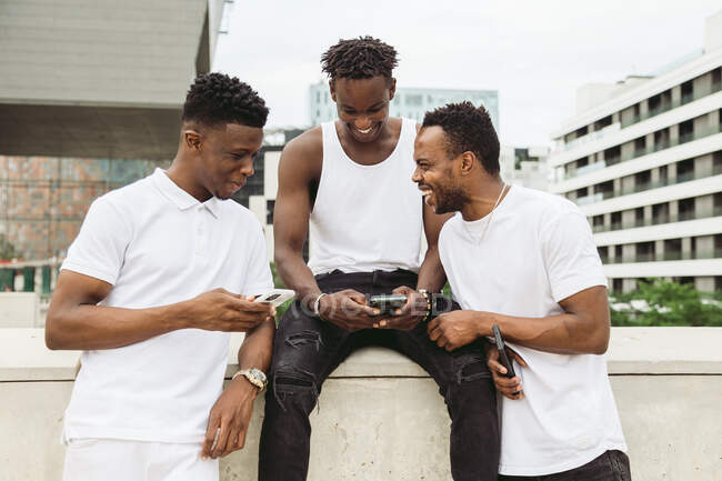 Amis masculins afro-américains positifs en vêtements décontractés partageant des téléphones cellulaires tout en riant joyeusement dans le parc — Photo de stock