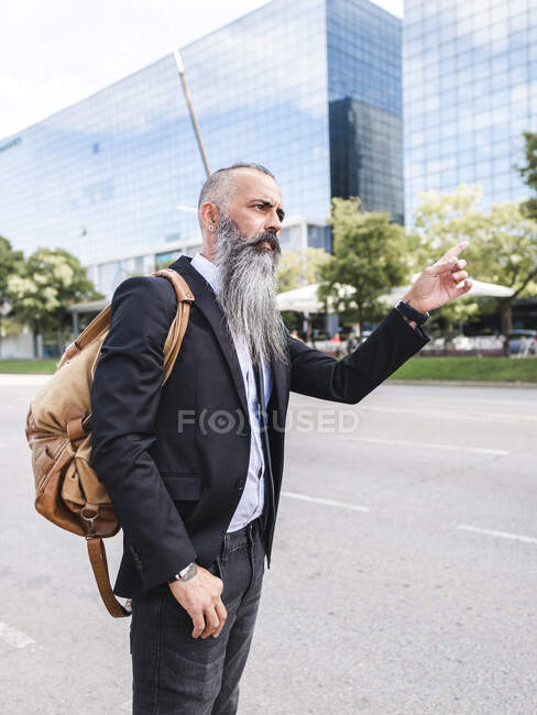 Вид збоку серйозного бородатого чоловіка в офіційному одязі, що стоїть поруч з дорогою, висікаючи таксі на вулиці з сучасними будівлями — стокове фото