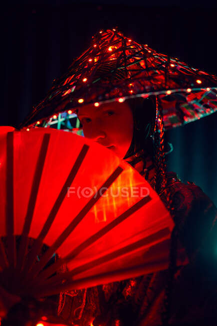 Жінка в автентичному вбранні та традиційному капелюсі зі світильниками, що стоять у темній студії з вентилятором у руці на чорному тлі — стокове фото