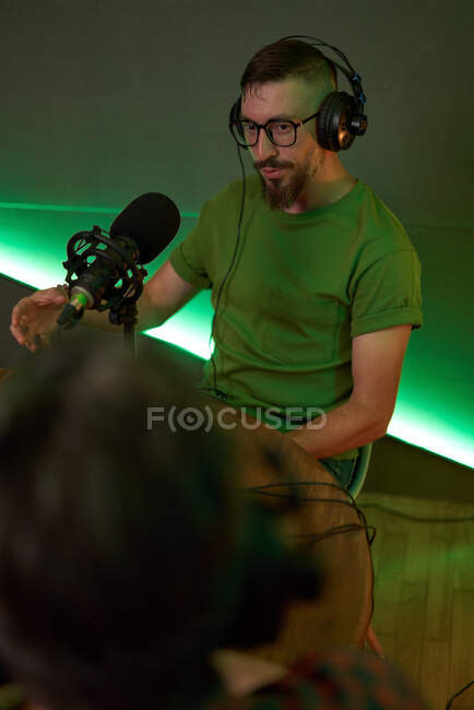 Junger männlicher Radiomoderator in lässiger Kleidung und Kopfhörer sitzt mit Mikrofon am Tisch und kommuniziert mit einem anonymen Kollegen während der Podcast-Aufzeichnung im Studio — Stockfoto