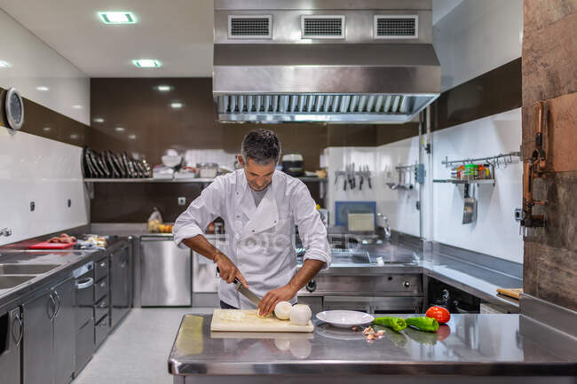 Chef masculino enfocado cortando mozzarella italiana con cuchillo mientras cocina comida en cocina del restaurante - foto de stock