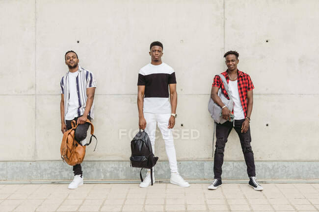 Corpo inteiro de amigos afro-americanos legais do sexo masculino com mochilas vestindo roupas da moda em pé perto do edifício e olhando para a câmera — Fotografia de Stock