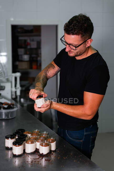 Стильный молодой татуированный мужчина-кондитер в повседневной одежде и очках закрывая крышку стеклянной банки со свежеприготовленным кремовым десертом в пекарне — стоковое фото