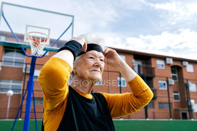 Mulher madura confiante em activewear e headband em pé perto de basquete aro em campo de esportes durante o treinamento na rua com a construção — Fotografia de Stock