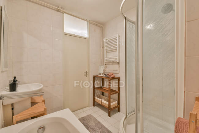 Interior bege claro de banheiro espaçoso vazio com cabine de chuveiro colocada perto da banheira e lavatório com produtos de higiene pessoal contra prateleiras de madeira com toalhas de banho — Fotografia de Stock