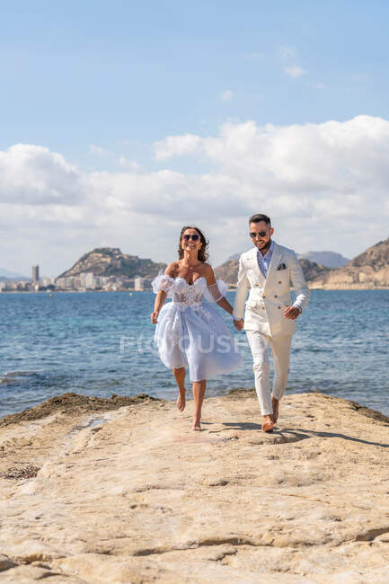 Полное тело веселой босой супружеской пары, бегущей по берегу у волнистого моря, наслаждаясь днем свадьбы в солнечной природе — стоковое фото