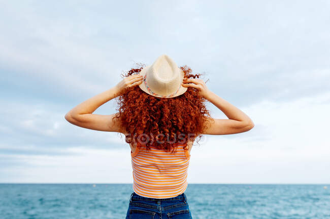 Vista trasera de hembra irreconocible con largos rizos de jengibre con sombrero de pie en la costa del mar azul - foto de stock