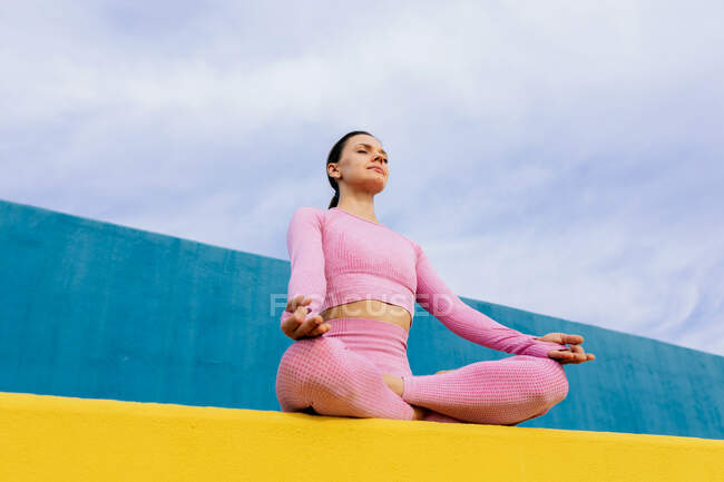 From below of young female in activewear sitting in lotus pose on sports ground while meditating — Stock Photo