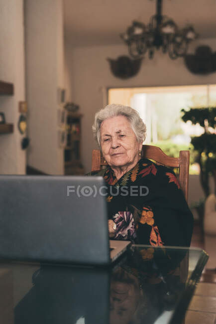 Mulher idosa com cabelos grisalhos curtos sentada na cadeira fazendo videochamada via netbook em casa — Fotografia de Stock