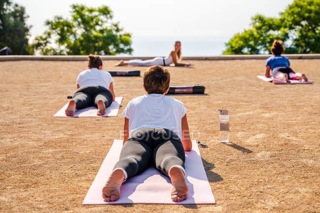 Rückansicht unkenntlich gemachter Frauen in aktiver Kleidung, die Salamba Bhujangasana liegen und ausführen, während sie im Freien Yoga im Park vor grünen Bäumen und wolkenlosem blauen Himmel bei sonnigem Tag praktizieren — Stockfoto