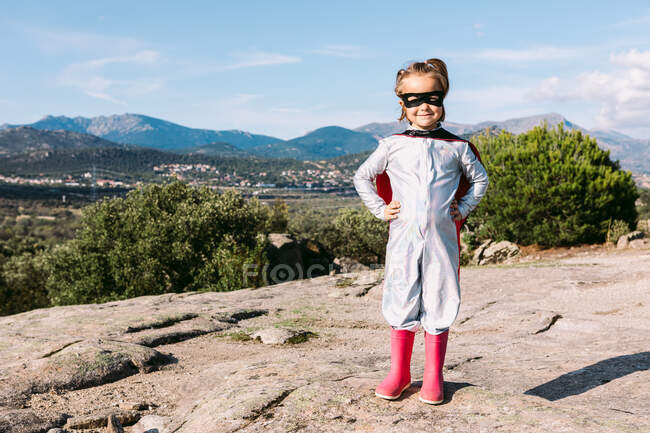 Полное тело маленькой девочки в костюме супергероя с руками на талии, стоящими на скалистом холме — стоковое фото