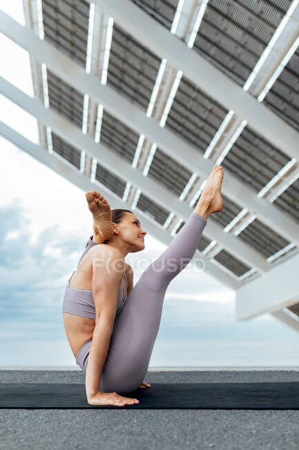 Вид збоку тіла тонкої жінки, що виконує позу йоги, сидячи на килимку з ногою за головою біля сонячної панелі — стокове фото