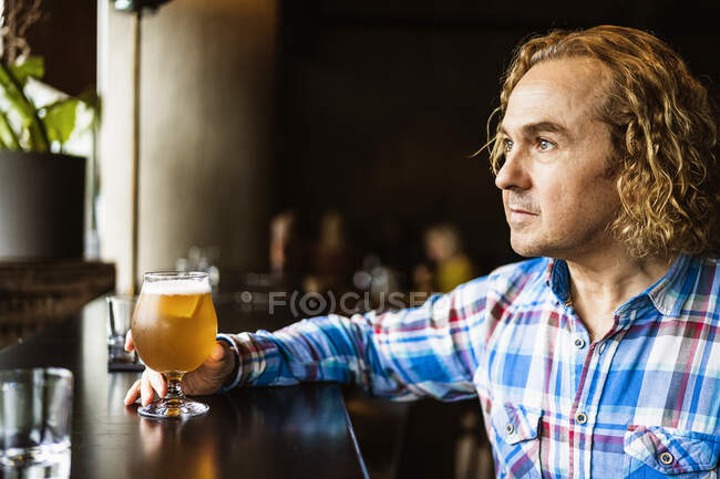 Thoughtful male with curly hair in casual clothes sitting at wooden counter near window in bar and drinking beer in daytime — Stock Photo