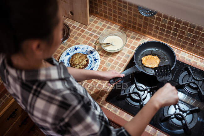 From above side view of focused young Asian female in casual clothes cooking yummy pancakes using spatula while standing in kitchen — Stock Photo