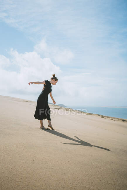Visão lateral de corpo inteiro de dançarina descalça estendendo os braços enquanto ela observa sua sombra lançada na areia — Fotografia de Stock