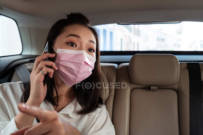 Pasajera étnica con máscara protectora sentada con cinturón de seguridad y dando instrucciones al taxista mientras está en una llamada telefónica - foto de stock