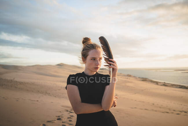 Femme sérieuse avec une plume noire portant une robe debout sur une dune de sable lavée par la mer au coucher du soleil — Photo de stock