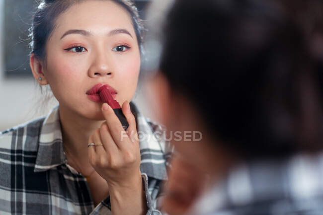 Молода приваблива азіатка в повсякденному одязі прикладає яскраву губну помаду, дивлячись на дзеркало. — стокове фото
