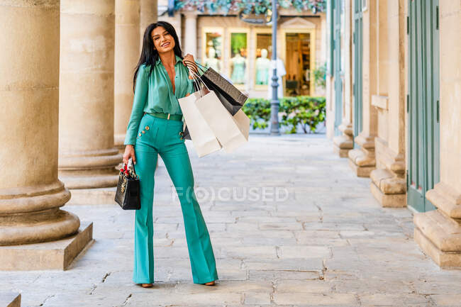 Full body of positive female in trendy outfit with shopping bags and purse looking at camera while standing near building with columns — Stock Photo