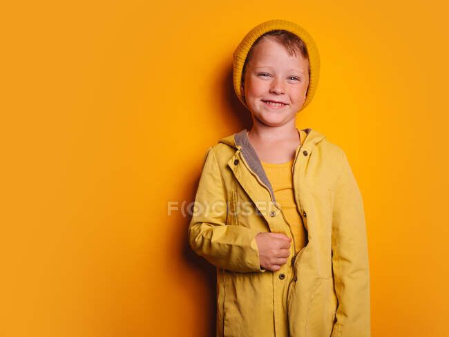Glücklicher Junge in leuchtend gelber Regenjacke und Mütze, lachend und vor gelbem Hintergrund im Studio in die Kamera blickend — Stockfoto