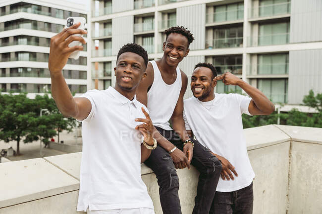 Positive African American man with friends showing victory gesture while fellow taking selfie on cellphone — Stock Photo