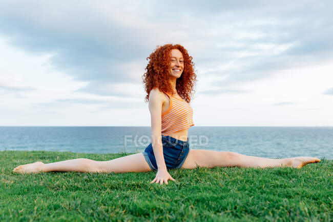 Delighted red haired female with curls doing Hanumanasana for stretching legs on grassy coast of rippling sea and looking at camera — Stock Photo