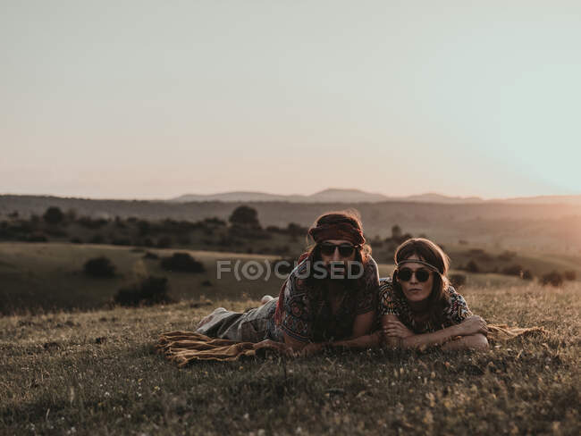 Hippie pareja con gafas de sol mirando a la cámara mientras se encuentra en el prado herboso en la naturaleza al atardecer - foto de stock