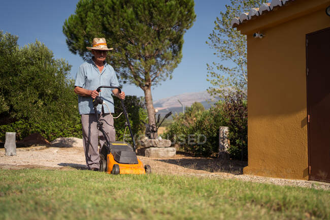 Full body of unrecognizable male gardener in hat mowing grassy lawn near bushes and trees in summer — Stock Photo