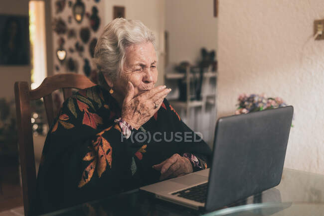 Mujer anciana positiva que usa ropa de abrigo sentada en la mesa y envía un beso de aire mientras habla por videollamada usando una computadora portátil - foto de stock
