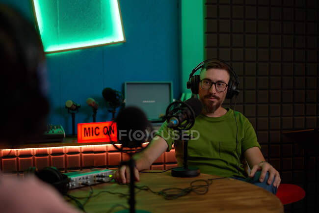 Positive young bearded male millennial in eyeglasses and headphones and talking in mic while recording podcast in dark studio — Stock Photo