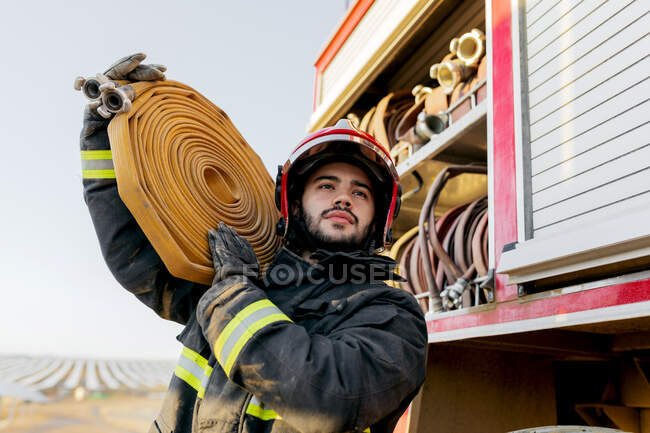 Von unten ein tapferer männlicher Feuerwehrmann mit Schutzhelm und Uniform, der wegschaut, während er einen großen schweren Schlauch auf der Schulter in der Nähe eines Feuerwehrfahrzeugs auf einem Acker trägt — Stockfoto