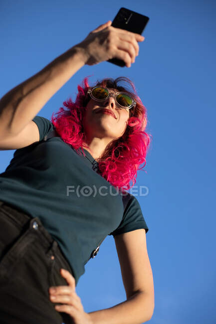 Desde abajo de la mujer confiada con el pelo rosa surfeando el teléfono celular mientras está de pie con la mano en la cintura contra el cielo azul claro - foto de stock
