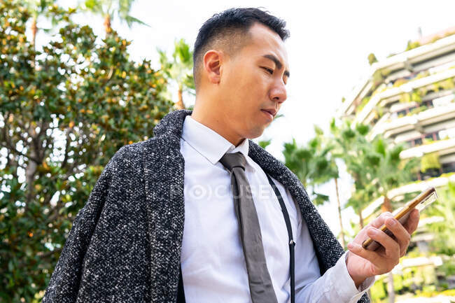 From below young ethnic male entrepreneur with tie looking at screen while speaking on cellphone in town — Stock Photo