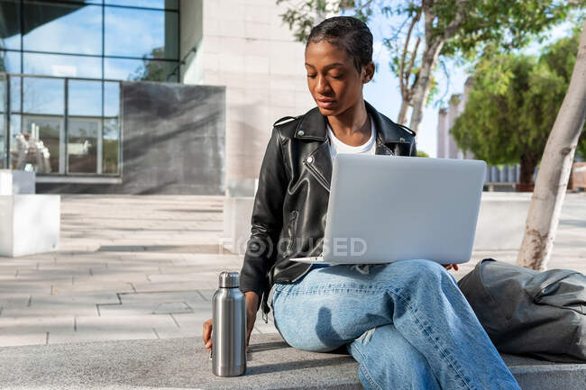 Contenuto Libera professionista afroamericana in giacca di pelle che prende thermos con bevanda calda mentre siede con netbook in grembo durante il lavoro online in strada — Foto stock