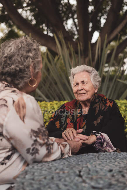 Mulher velha sorrindo e de mãos dadas enquanto se senta à mesa no quintal exótico com palmas e arbustos e desfrutando de conversa com idosas amigas — Fotografia de Stock