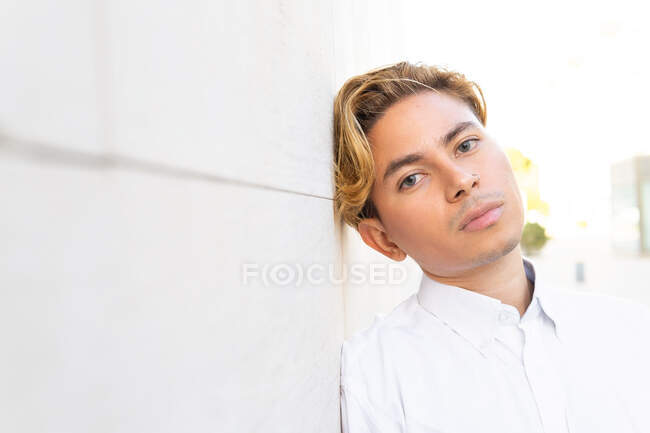 Serious young male in white shirt looking at camera with thoughtful gaze while leaning on wall on street in city — Stock Photo