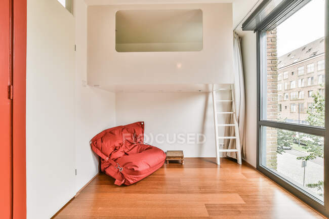 Silla de bolsa suave roja colocada en la esquina de la habitación vacía con escalera blanca cerca de la ventana panorámica de vidrio en el apartamento moderno - foto de stock