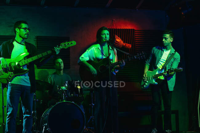 Group of people in casual clothes playing guitars and drums while woman singing and performing song in club with neon lights — Stock Photo