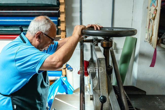Seitenansicht des fokussierten älteren Handwerkers in Freizeitkleidung und Schürzenspinnrad während der Bedienung der Bindemaschine in der Druckerei — Stockfoto