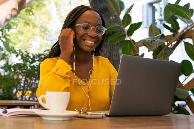 Fröhliche Afroamerikanerin in Kopfhörern mit Videoanruf auf Laptop, während sie mit einer Tasse Kaffee am Tisch in der Cafeteria im Freien sitzt — Stockfoto