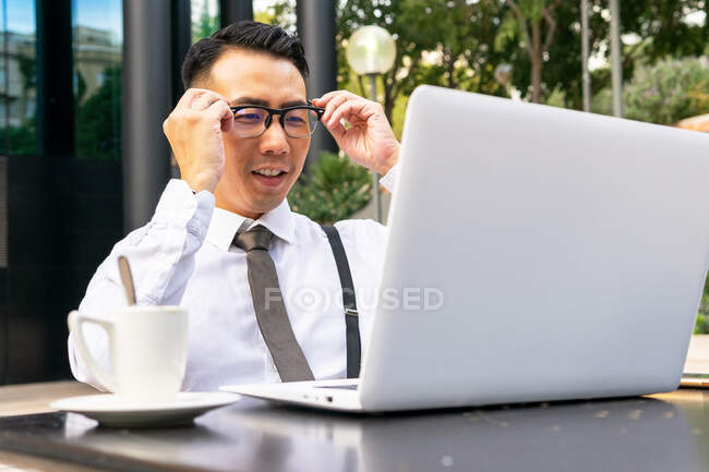 Gut gekleideter männlicher Jungunternehmer setzt Brille gegen Tisch mit Netbook und Heißgetränk in Straßencafeteria auf — Stockfoto